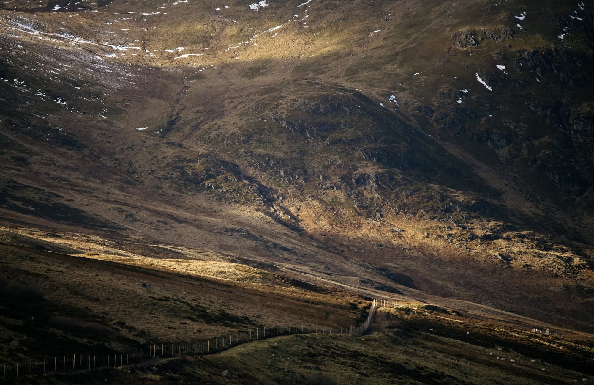 A colour photograph of a large, rugged looking hill with a small fences off path runnning across the bottom.