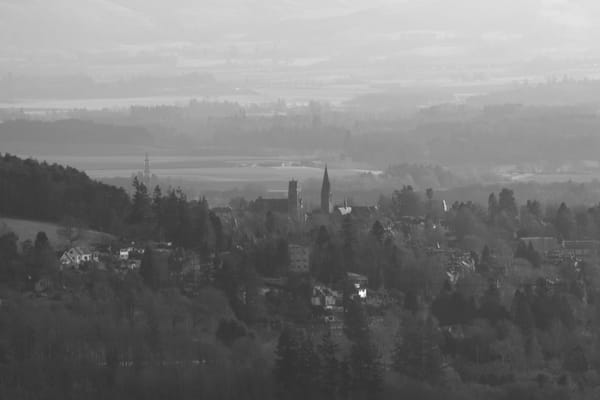 A black and white photograph of a town set within lots of trees from a distance. You can see church spires and houses.